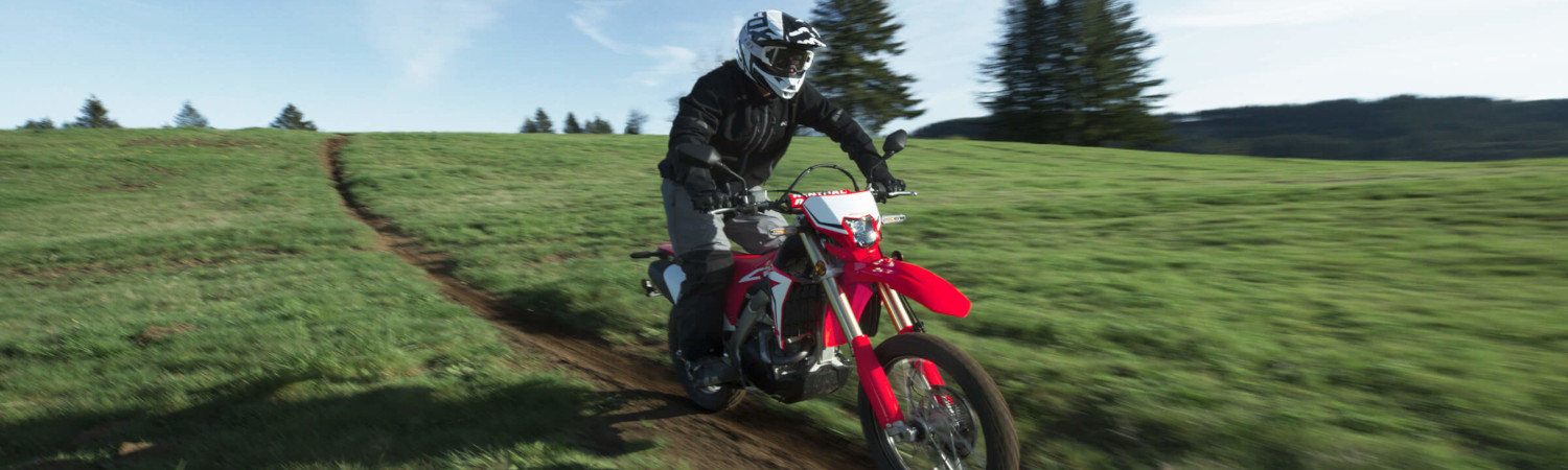 A person riding a red Honda® motorcycle on a dirt trail through a field on a sunny day.